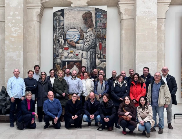 Groupe de personnes posant devant une grande mosaïque représentant un scientifique dans un bâtiment en pierre.